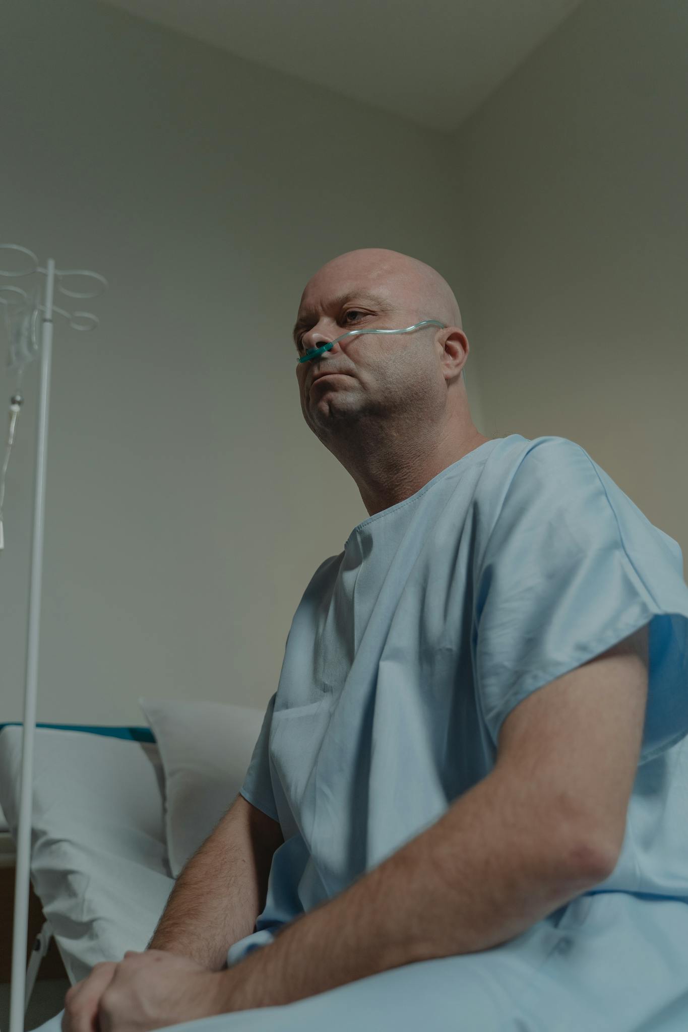 Bald man sitting in hospital room wearing a gown and nasal cannula, depicting illness.
